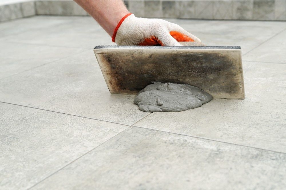 A Gloved Hand Uses a Trowel to Spread Gray Grout Between Light-coloured Tiles on a Floor — North Coast Tiles in Maclean, NSW