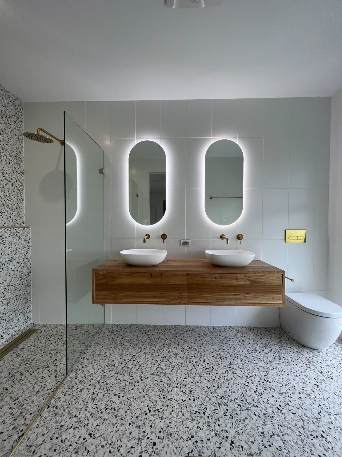 Modern bathroom with floating wood vanity, two oval backlit mirrors, terrazzo floor, and a glass shower — North Coast Tiles in Grafton, NSW