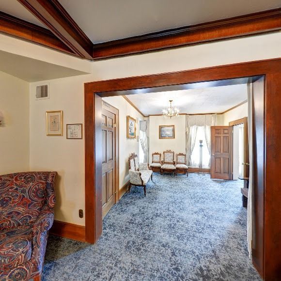Interior view of a room with wood trim and blue carpet, leading to another room with chairs and a chandelier.