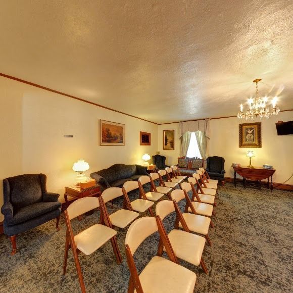 Funeral home interior with chairs, sofa, and artwork. Beige walls, carpet, and chandelier.
