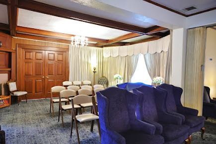 A funeral home chapel with chairs set for a service. Wooden doors, neutral curtains, and a chandelier.