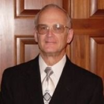 Man in suit and glasses smiles, standing in front of a wooden door.