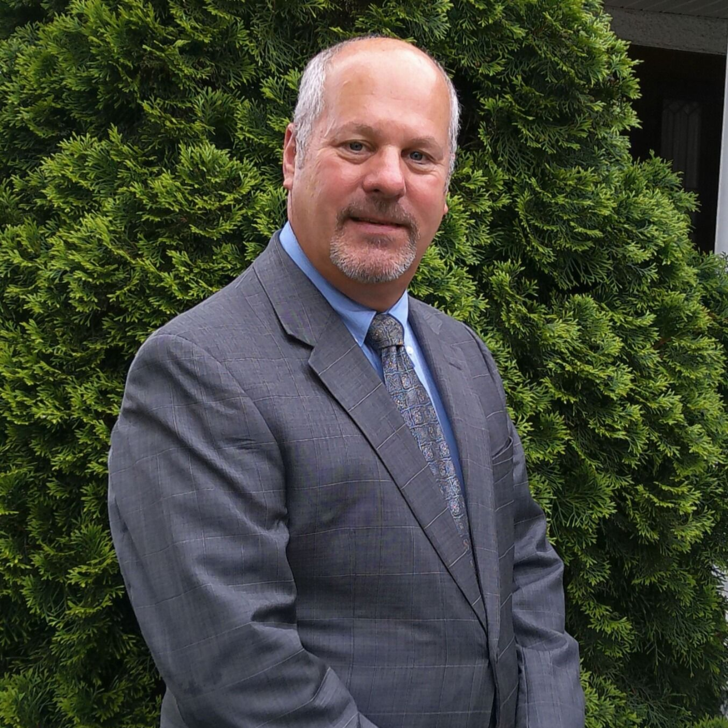 Man in a gray suit and tie, smiling, standing in front of a green bush.