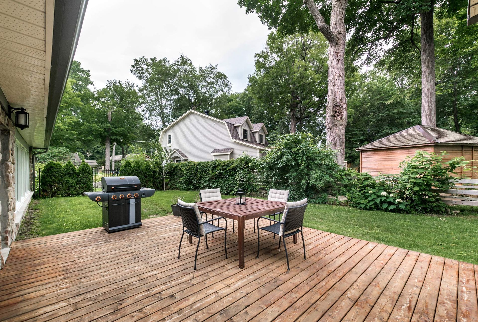 Backyard patio with a wooden deck, table and chairs, grill, and surrounding trees and greenery. A house is in the background.