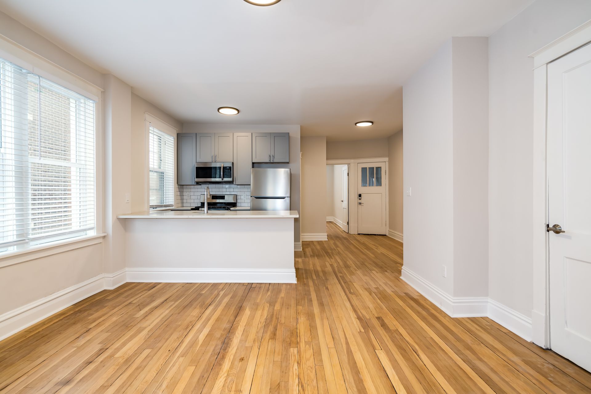 An empty living room with hardwood floors and a kitchen in the background.