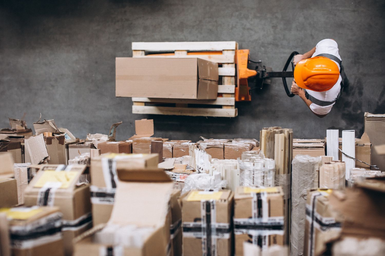 Warehouse worker in orange hard hat operating a pallet jack loaded with boxes. Many other boxes are in foreground.