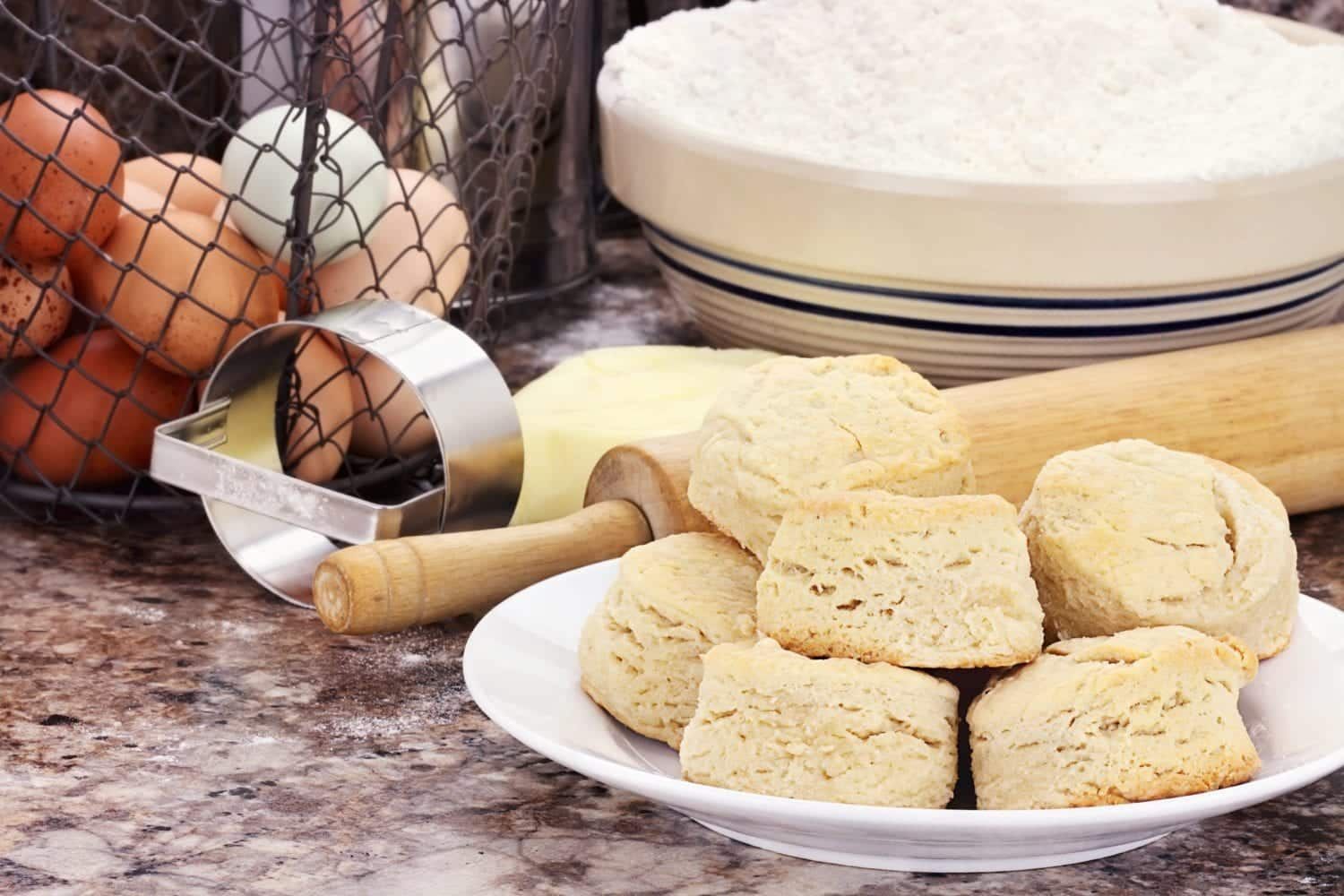 A plate of biscuits is sitting on a counter next to eggs and a rolling pin.