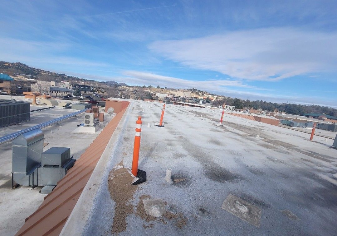 Overhead view of a flat, snowy roof with orange safety poles, with buildings and a bright sky in the background.