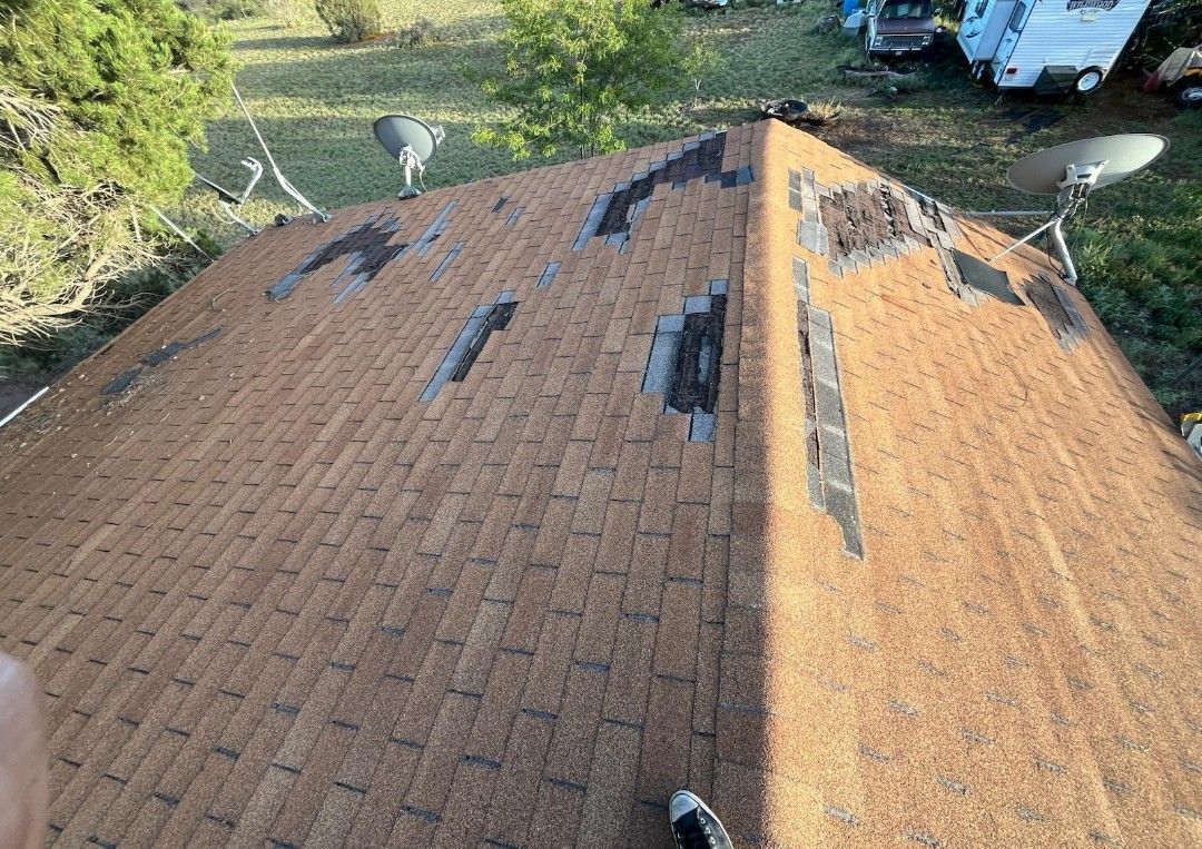 Damaged brown shingled roof with multiple holes, viewed from above, surrounded by grass and trees.