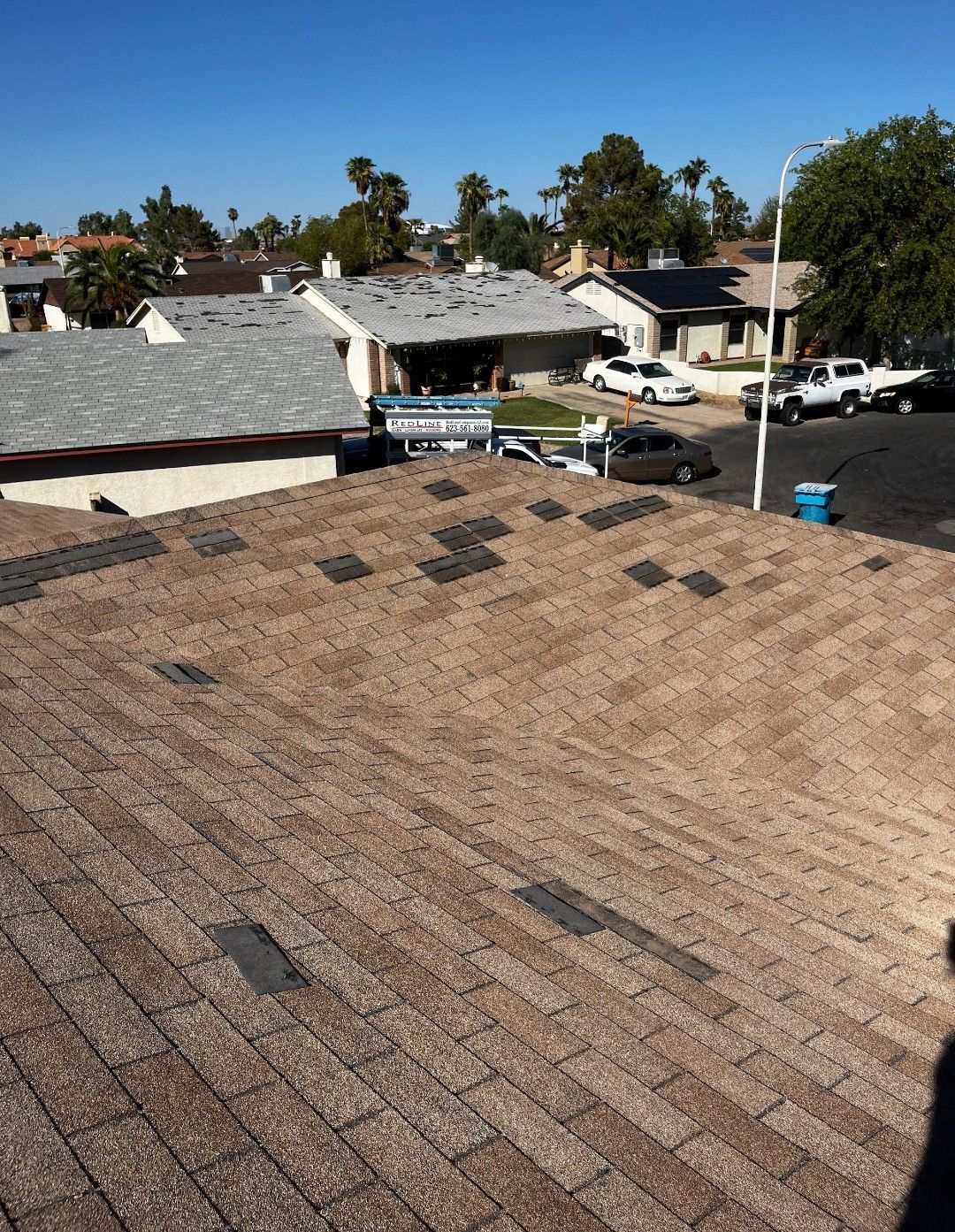 Brown shingle roof with missing and damaged shingles, houses in background, sunny day.