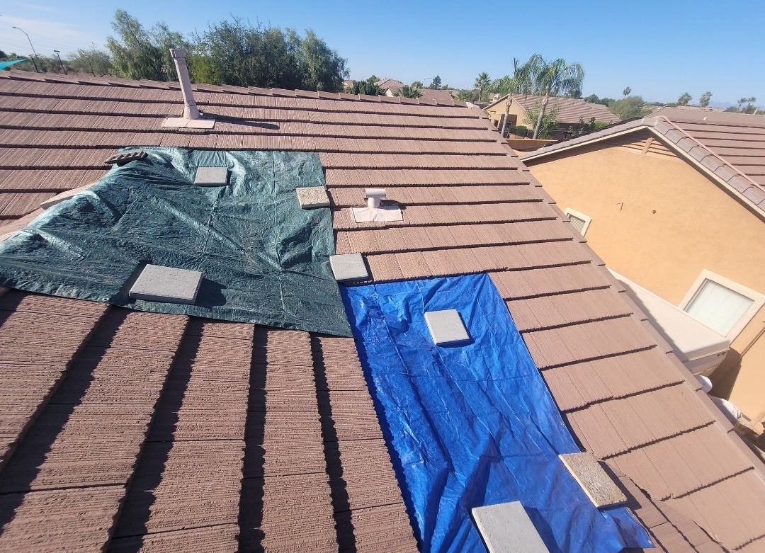 Brown tile roof with blue and green tarps, concrete blocks, and vents under a blue sky.