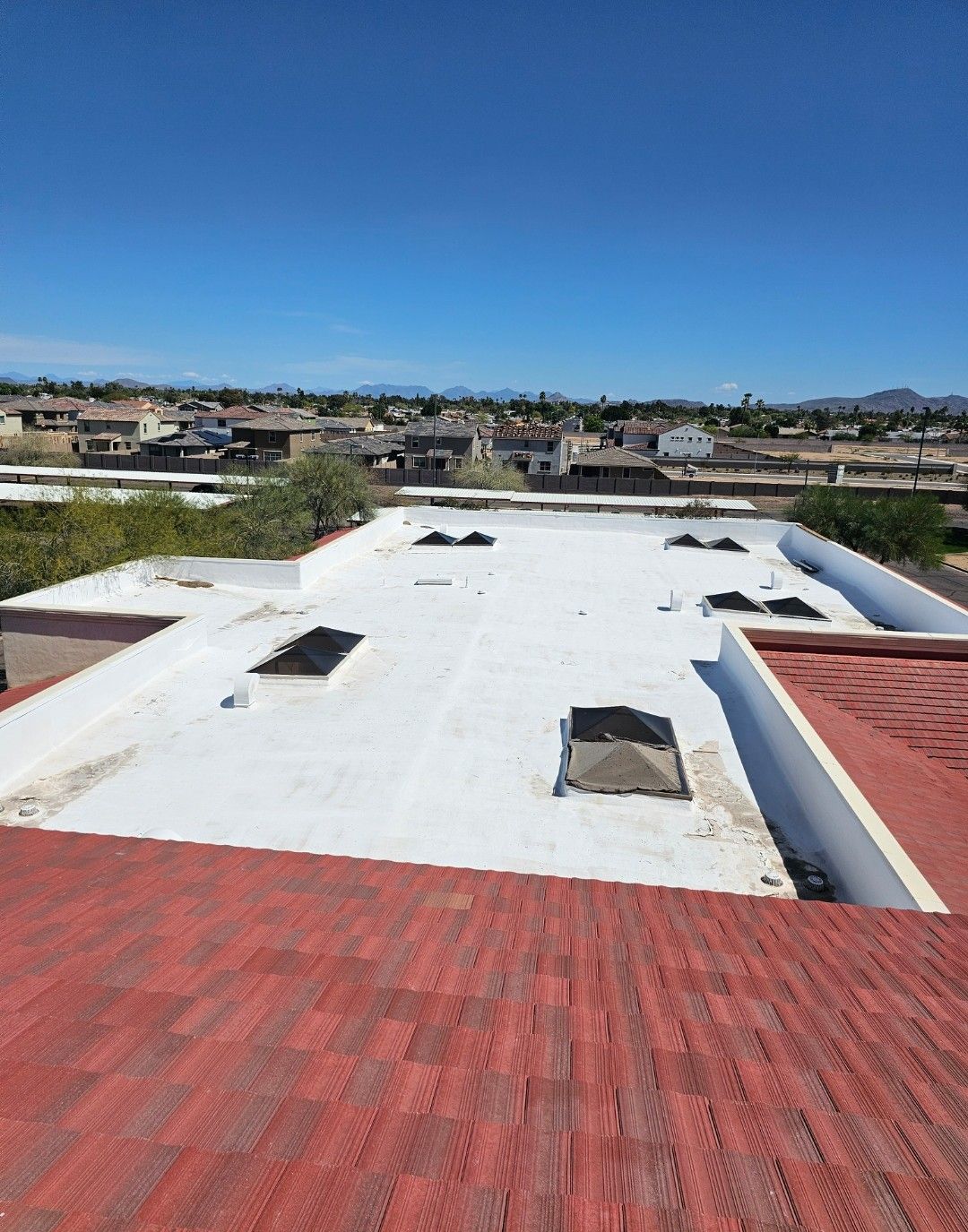 View of white flat roof with black vents, red tile roof, buildings and blue sky.