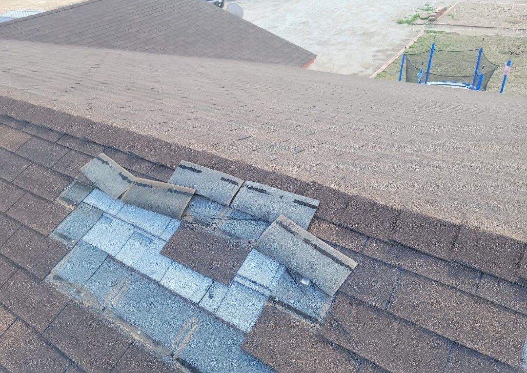 Damaged asphalt roof shingles with missing and lifted sections, with a brown color palette.