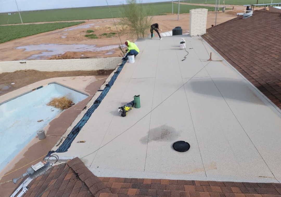Roofers working on a flat roof. One worker applies sealant, others stand nearby. Brown shingles border the roof.