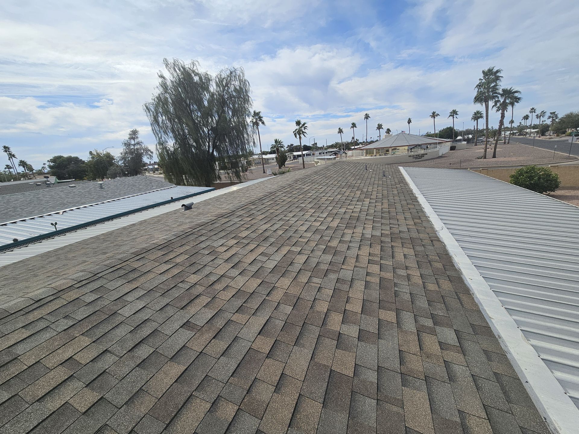 Roof with brown shingles, blue sky, palm trees in the distance.