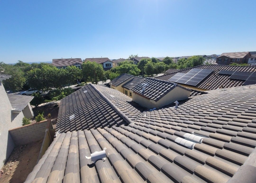 Rooftop view of multiple houses with tile roofs; solar panels on one, trees, and blue sky.