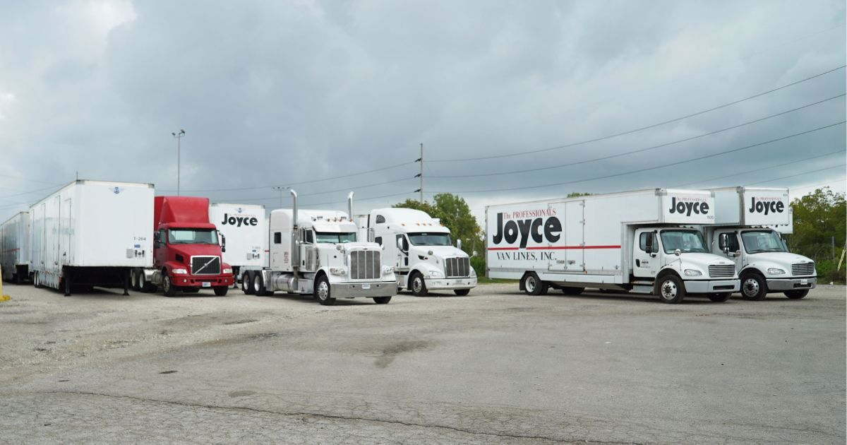 Delivery trucks and trailers parked in a gravel lot under a cloudy sky.