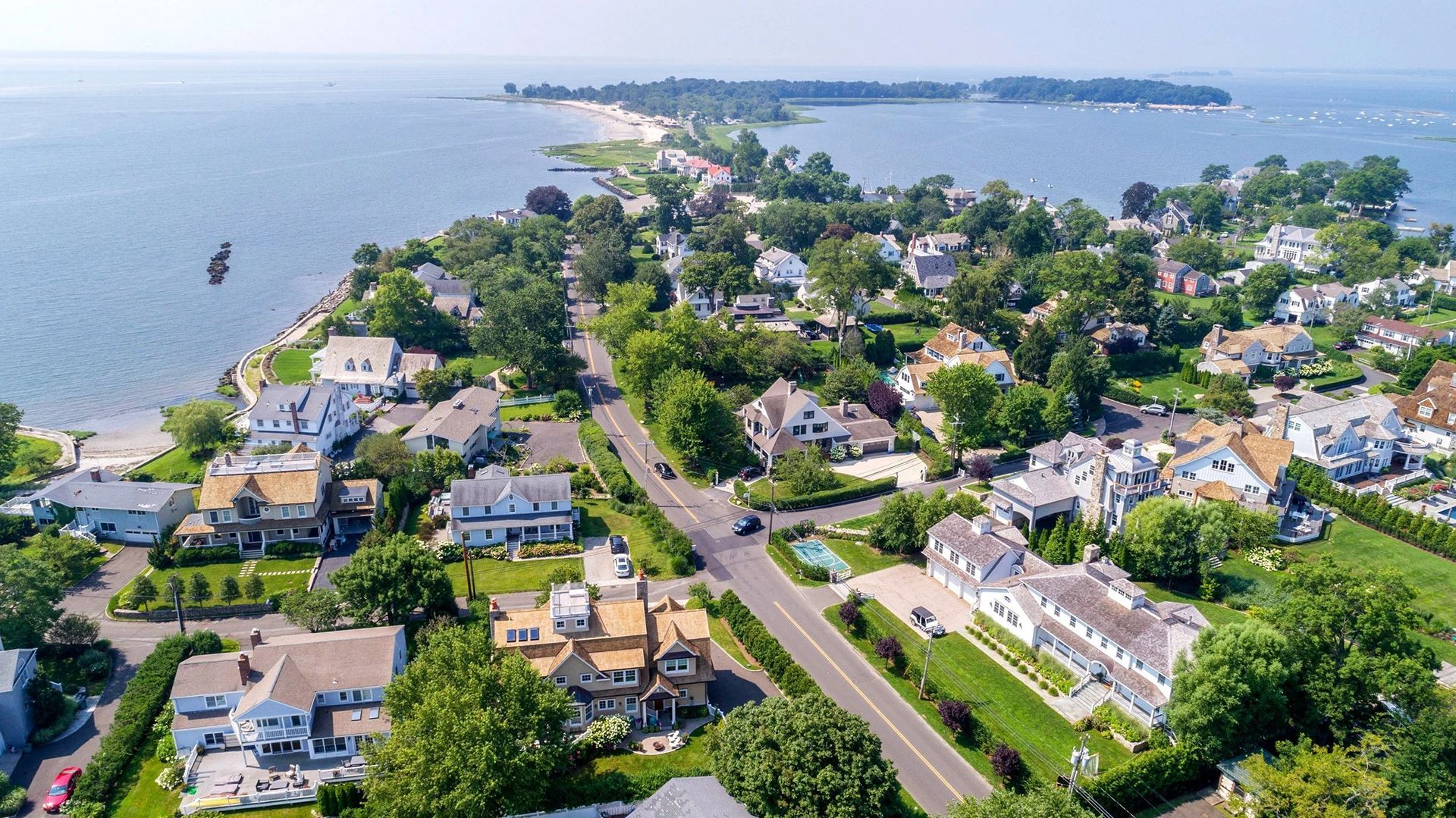 Aerial view of a coastal neighborhood with houses, a road, and a sandy peninsula extending into the ocean.