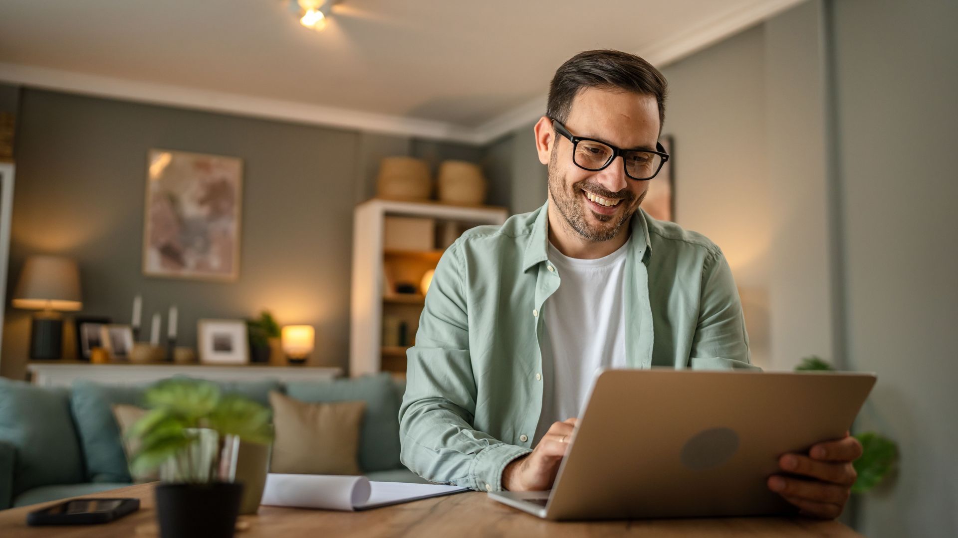Smiling man in glasses works on a laptop at a desk in a cozy living room.