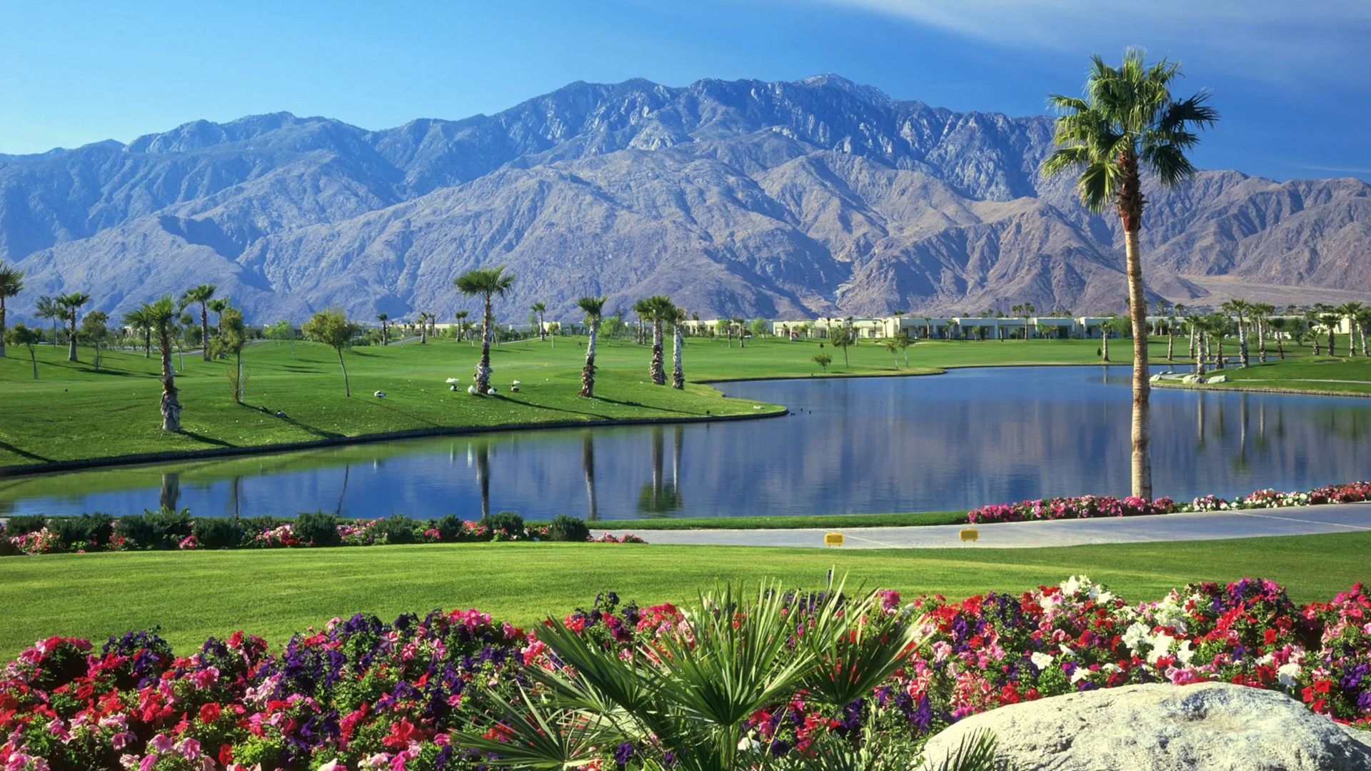 Lush green golf course with lake, palm trees, and mountains in the background under a blue sky.