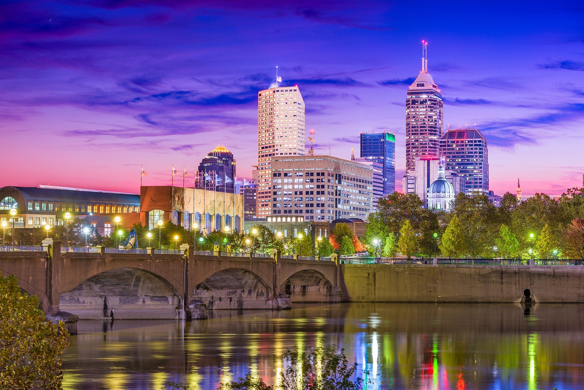 Skyline of Indianapolis at dusk, buildings reflected in the canal water under a purple and pink sky.