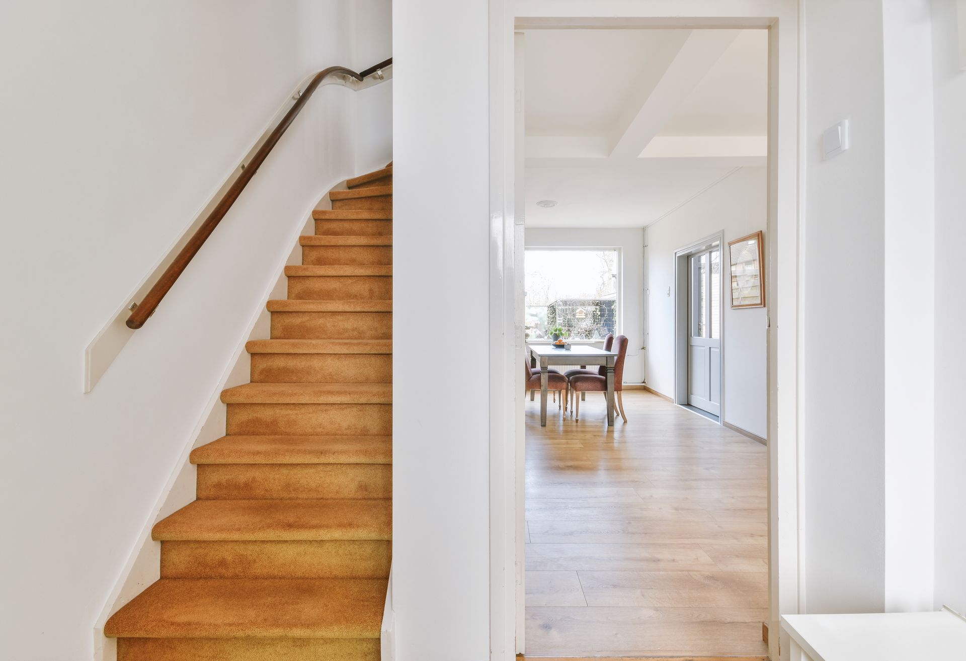 Staircase with carpet leads up. View through doorway to dining room with wooden floor.