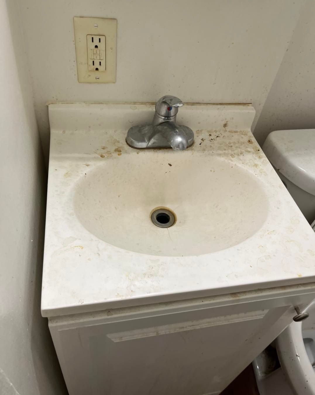 Dirty white bathroom sink with chrome faucet, electrical outlet above.