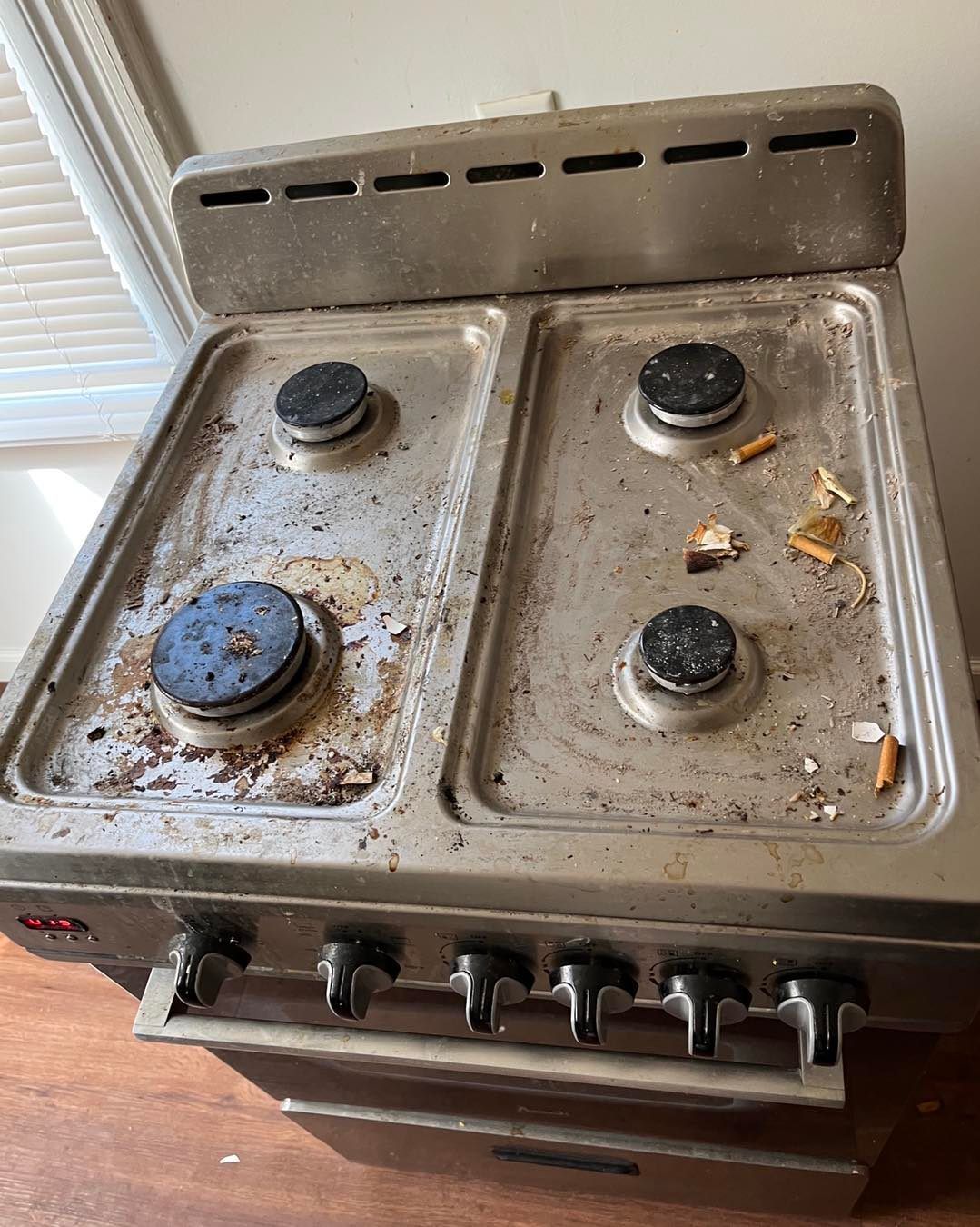 Dirty, stainless steel stovetop with four burners, food debris, and cigarette butts.