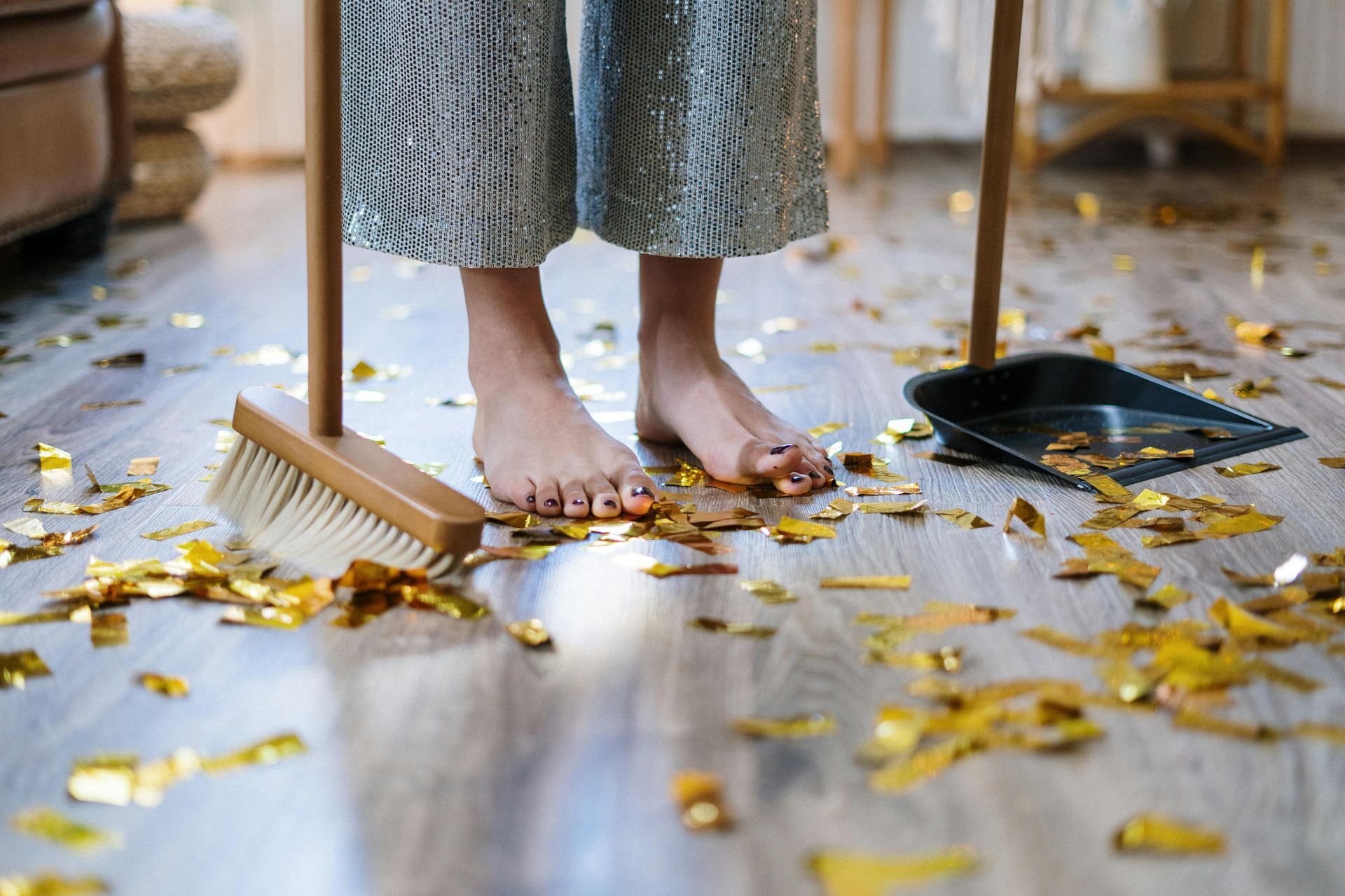 Bare feet sweeping gold confetti with a broom and dustpan on a wooden floor.