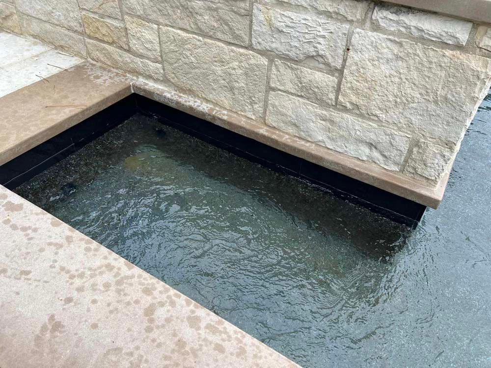 Rectangular water feature with stone walls; water overflowing from the contained basin into a surrounding pool.