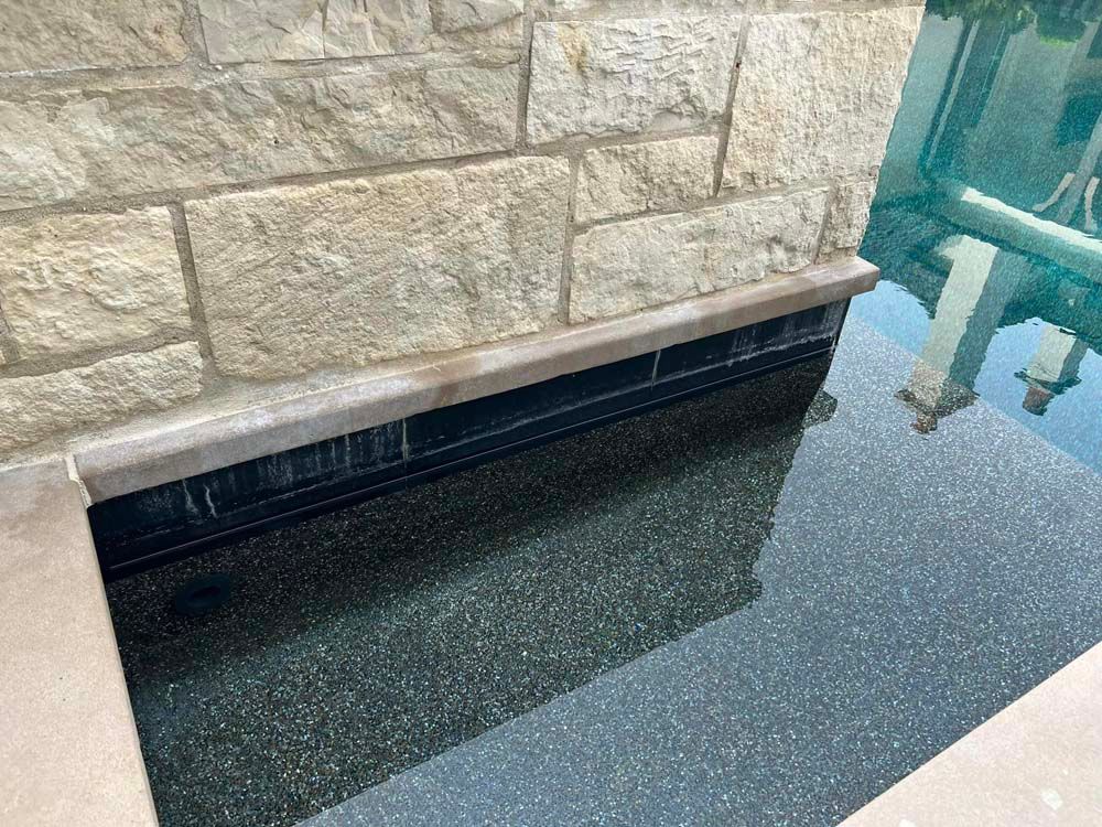 Stone wall with a spillway into a dark-tiled pool; water cascades over the edge.