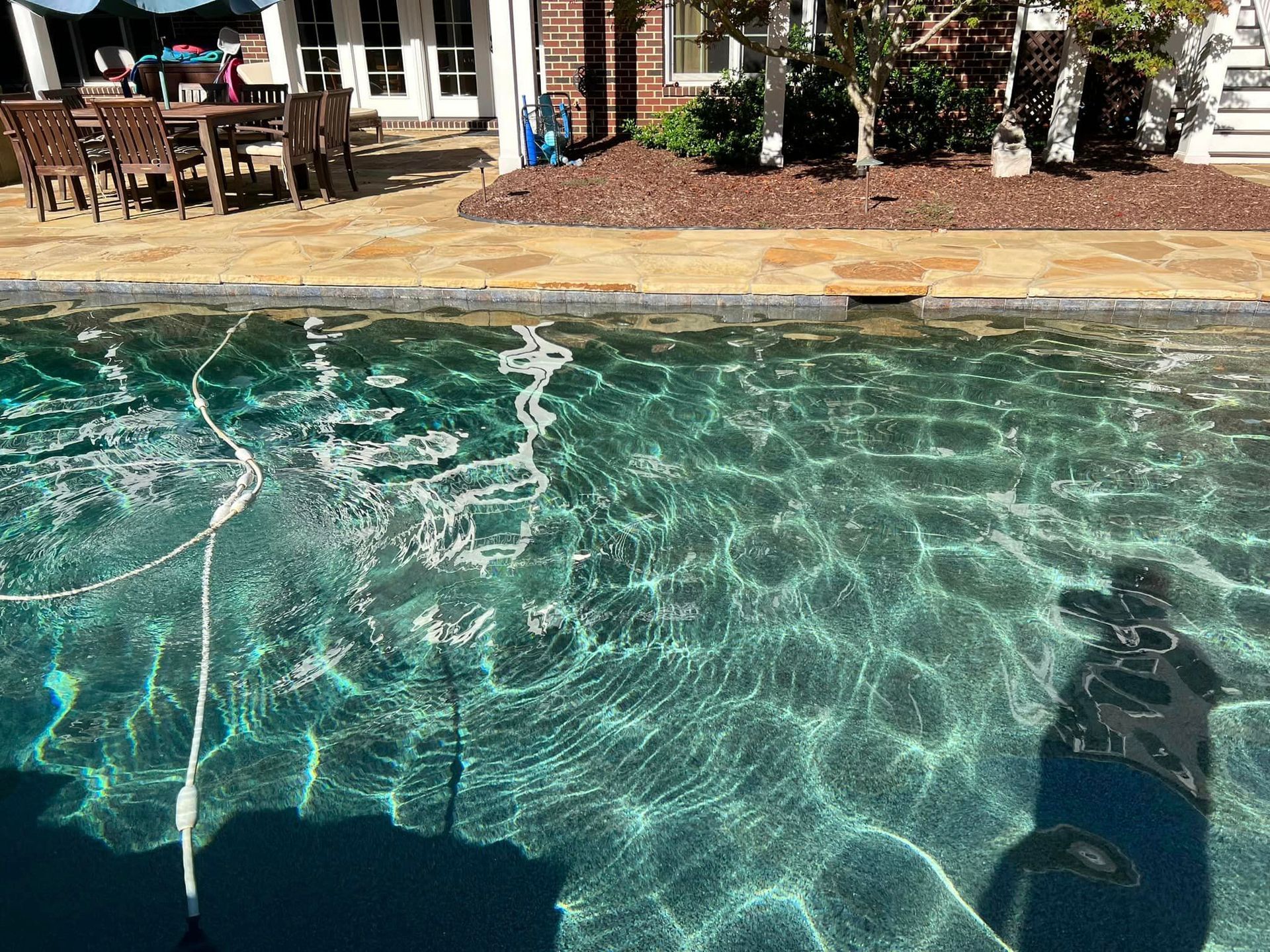 Pool with rippling turquoise water, stone coping, and a backyard with furniture visible in the background.