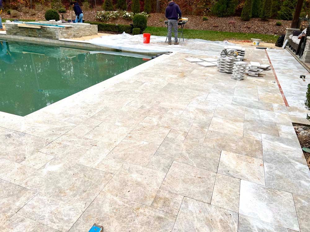 Workers tiling around a pool. Beige stone tiles, green pool water, and a grassy backyard are visible.