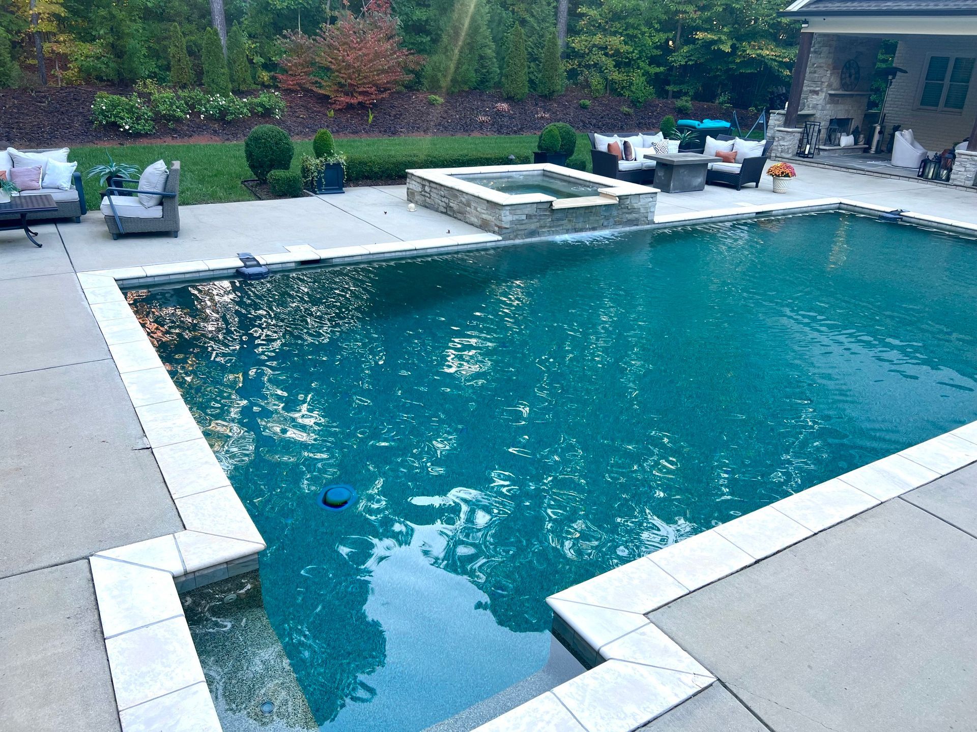 Pool with surrounding patio, hot tub, and outdoor seating in a backyard. Blue water, gray concrete, greenery.