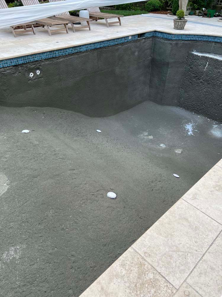 Empty swimming pool with rough, gray plaster interior, surrounded by light-colored stone and blue-tiled coping.