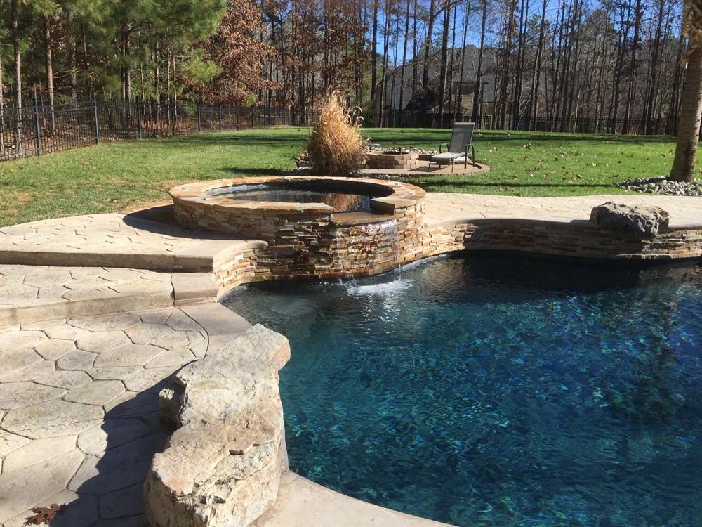 Pool with dark blue water and a stone waterfall feature, next to a stone hot tub. Green grass in the background.