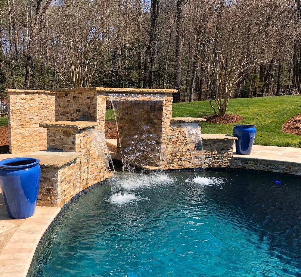 Swimming pool with stone waterfall feature; blue pots, green grass, and trees in the background.