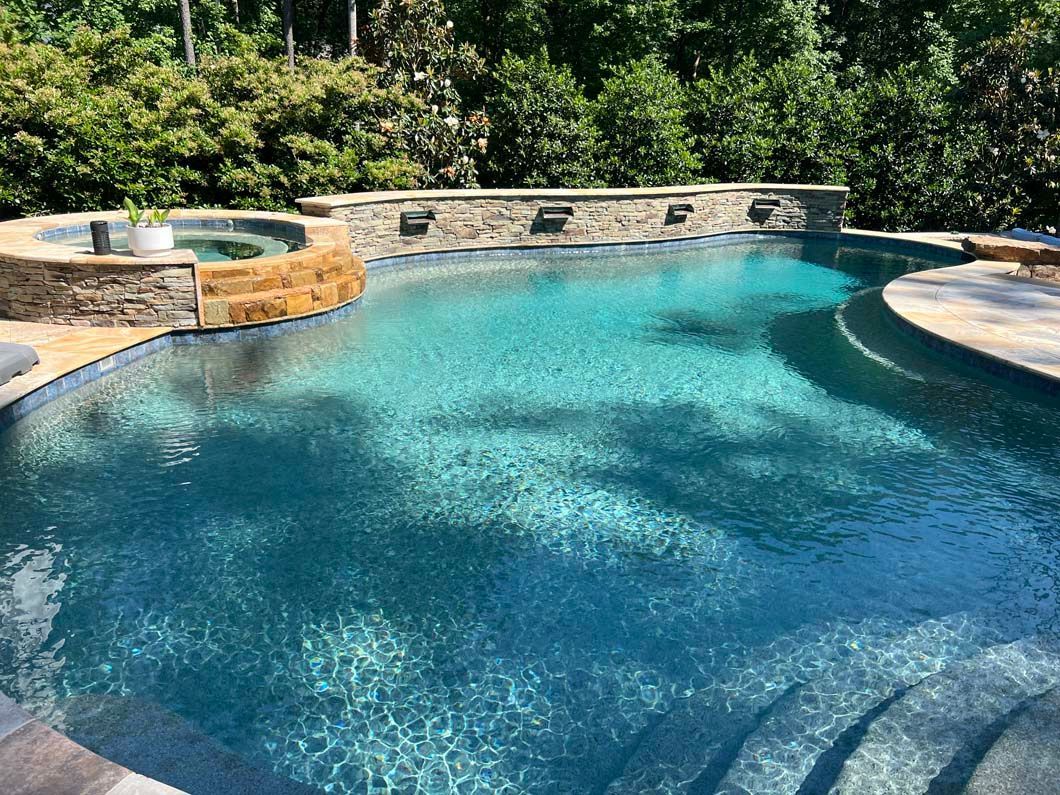 Pool with turquoise water and stone accents, a built-in hot tub, and lush green trees in the background.