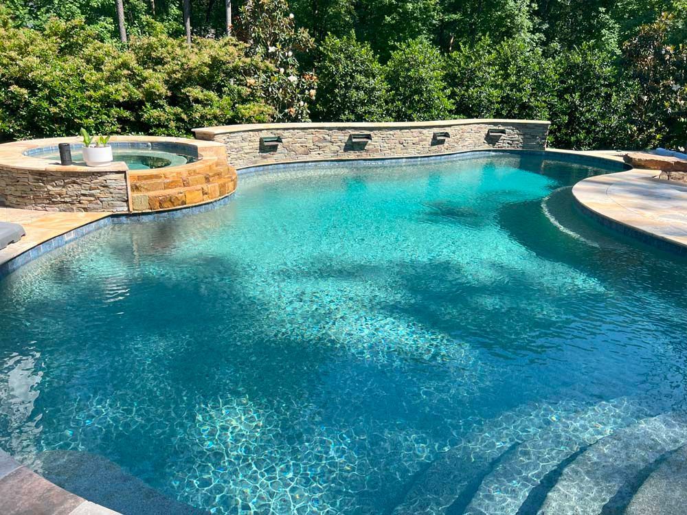 A turquoise-colored swimming pool with a connected jacuzzi and a stone wall with water features.