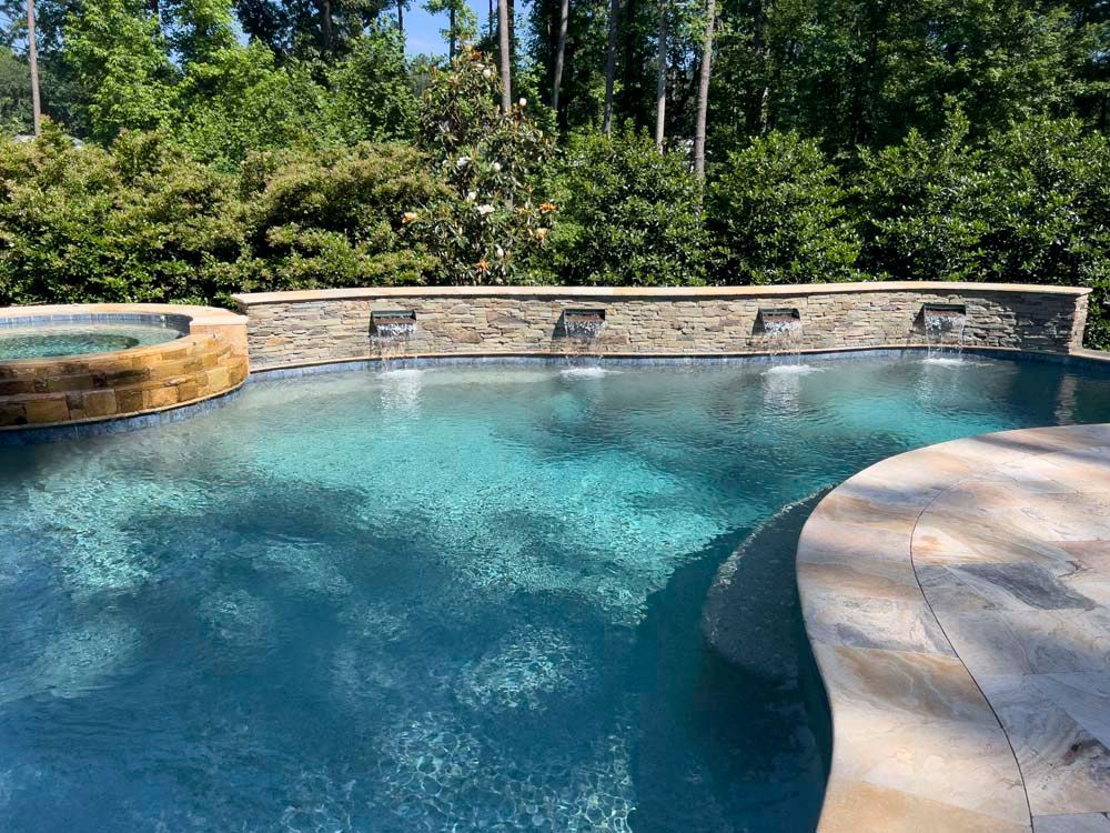 Swimming pool with water features, surrounded by stone and greenery. Blue water, sunny day.