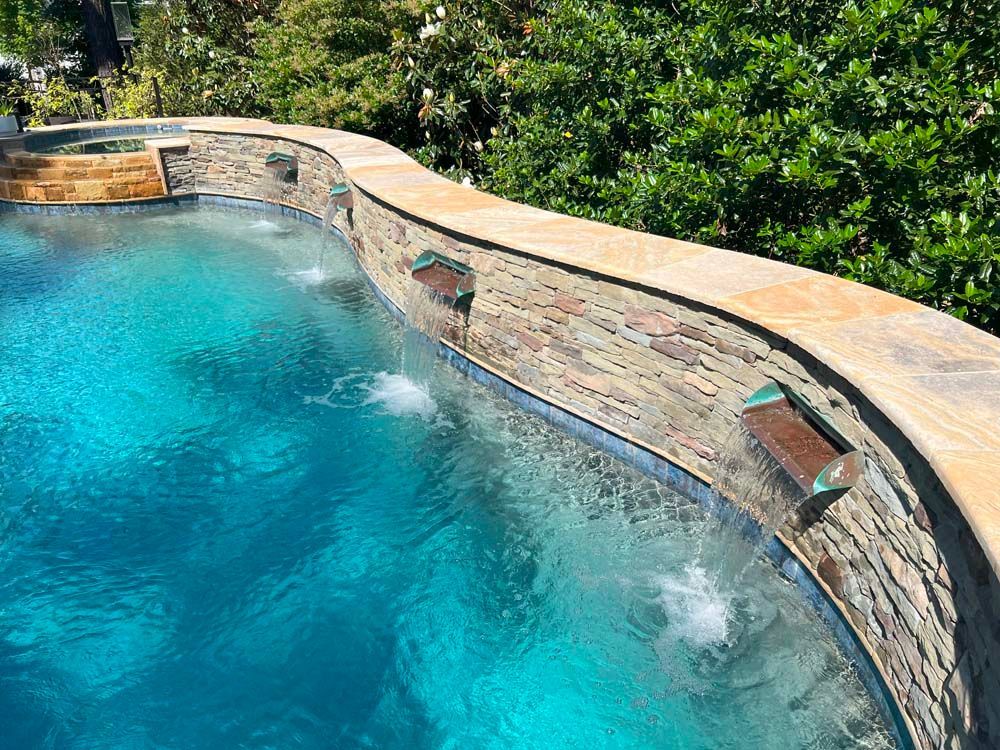 Curving pool with blue water, stone wall with copper water features, lush greenery in background.
