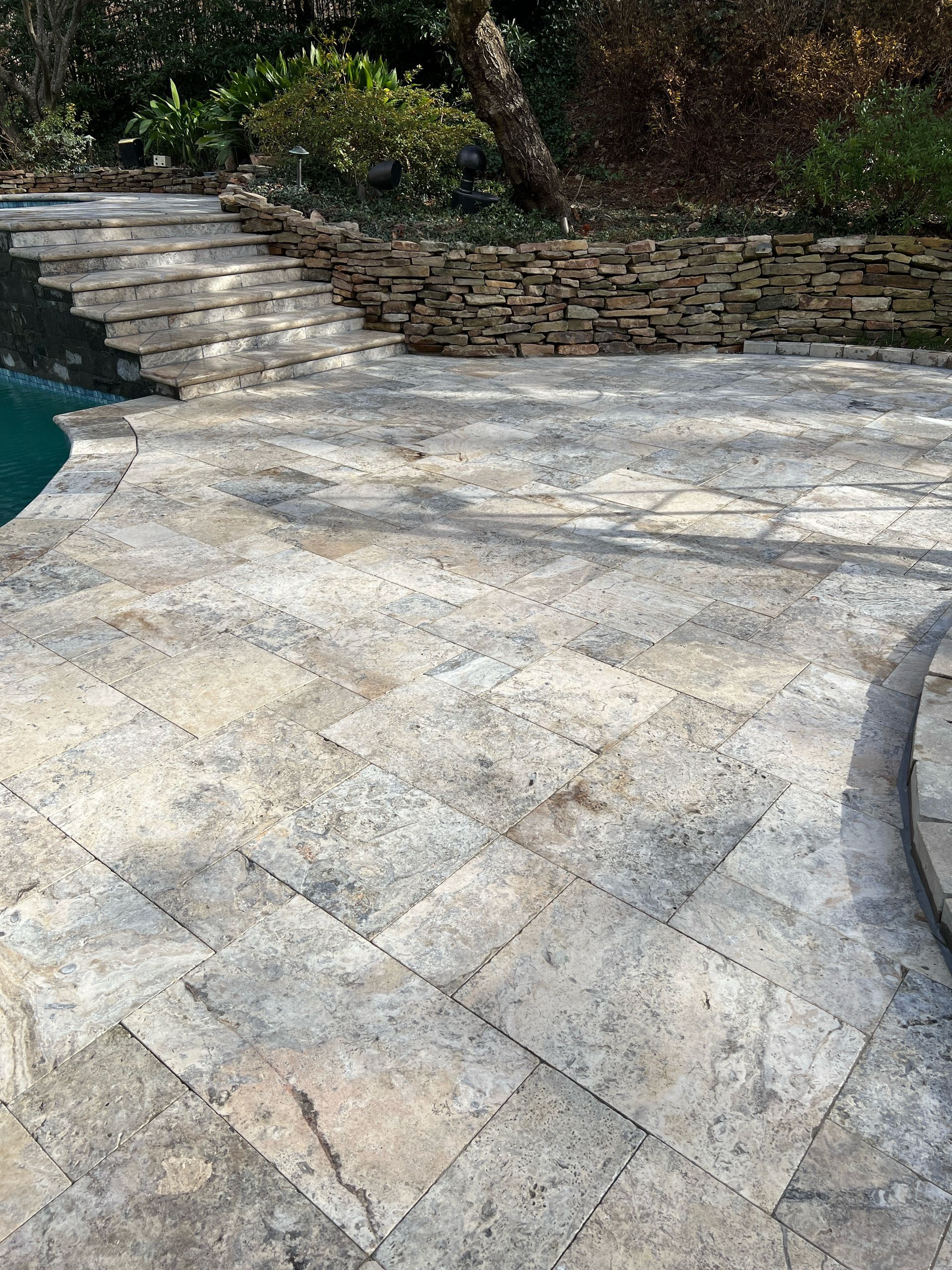 A patio with a swimming pool in the background and a stone wall.