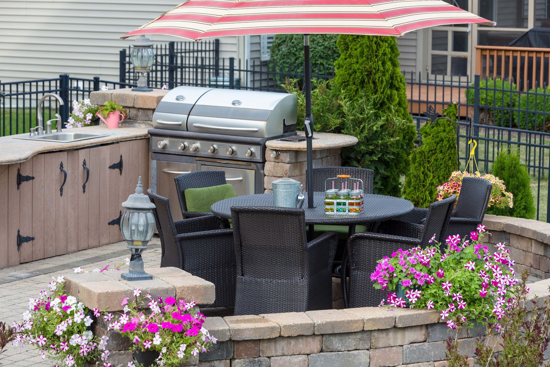 Outdoor patio with grill, dining table, and seating under a striped umbrella, surrounded by flower planters and a black fence.