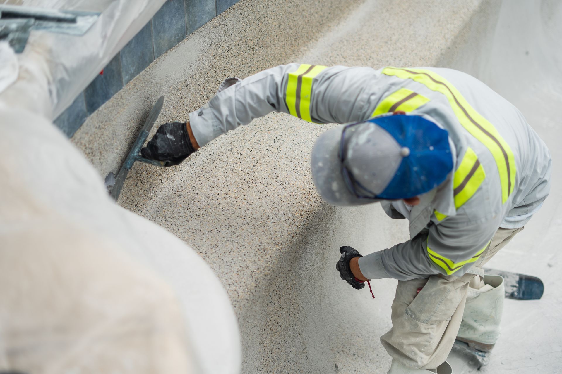 Person applying plaster to a pool wall with a trowel, wearing a blue cap, and safety vest.