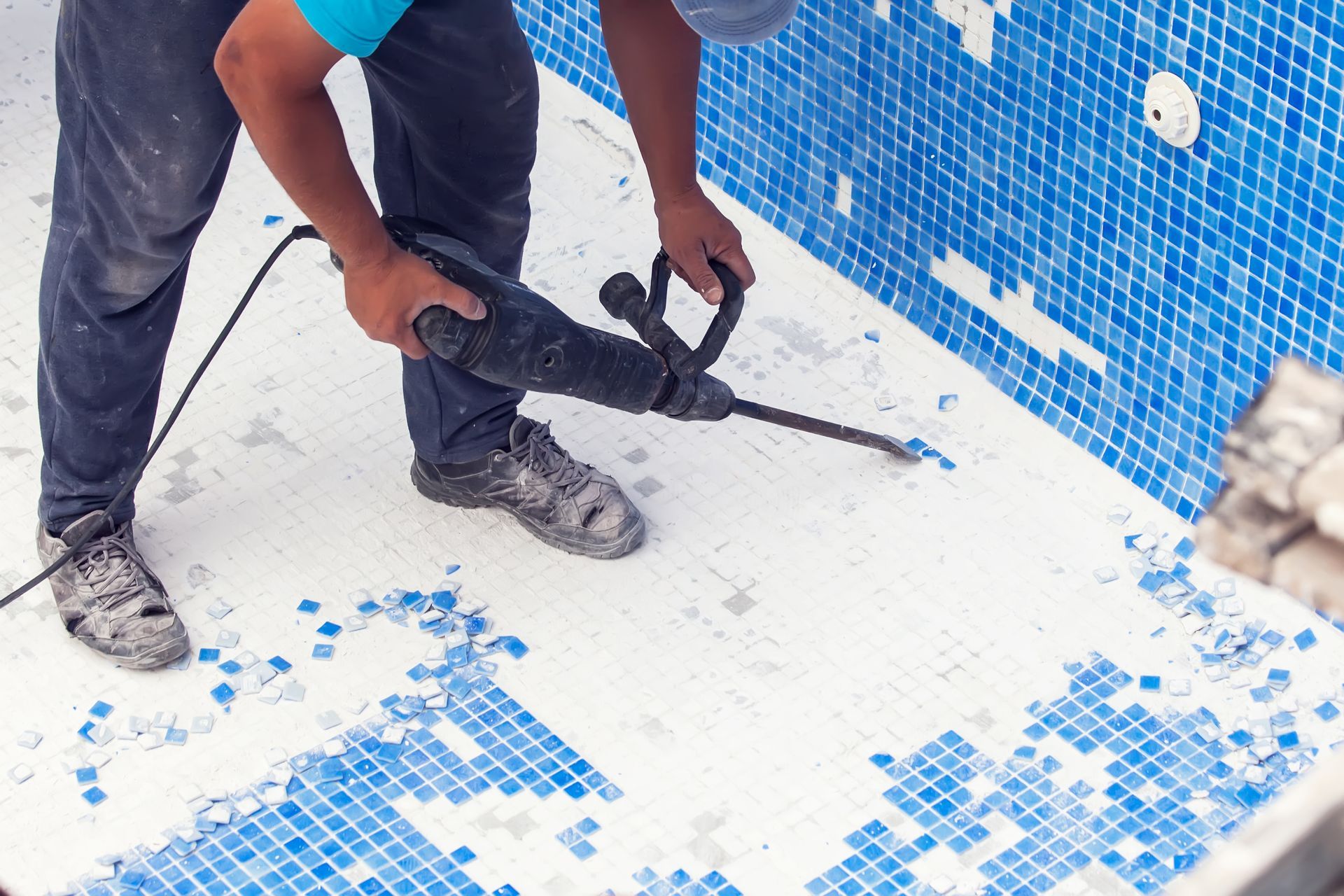 Person using a jackhammer to remove blue mosaic tiles from a swimming pool.