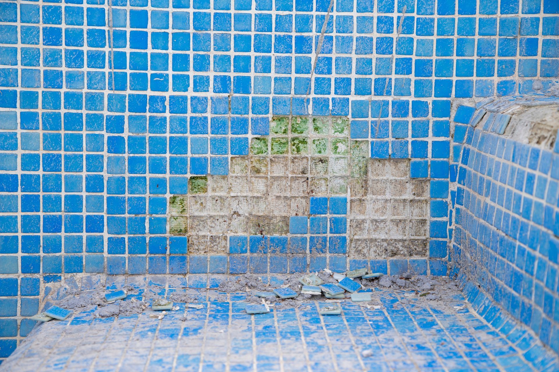 Damaged blue tile in a pool; missing tiles reveal concrete beneath. Debris lies on the pool floor.