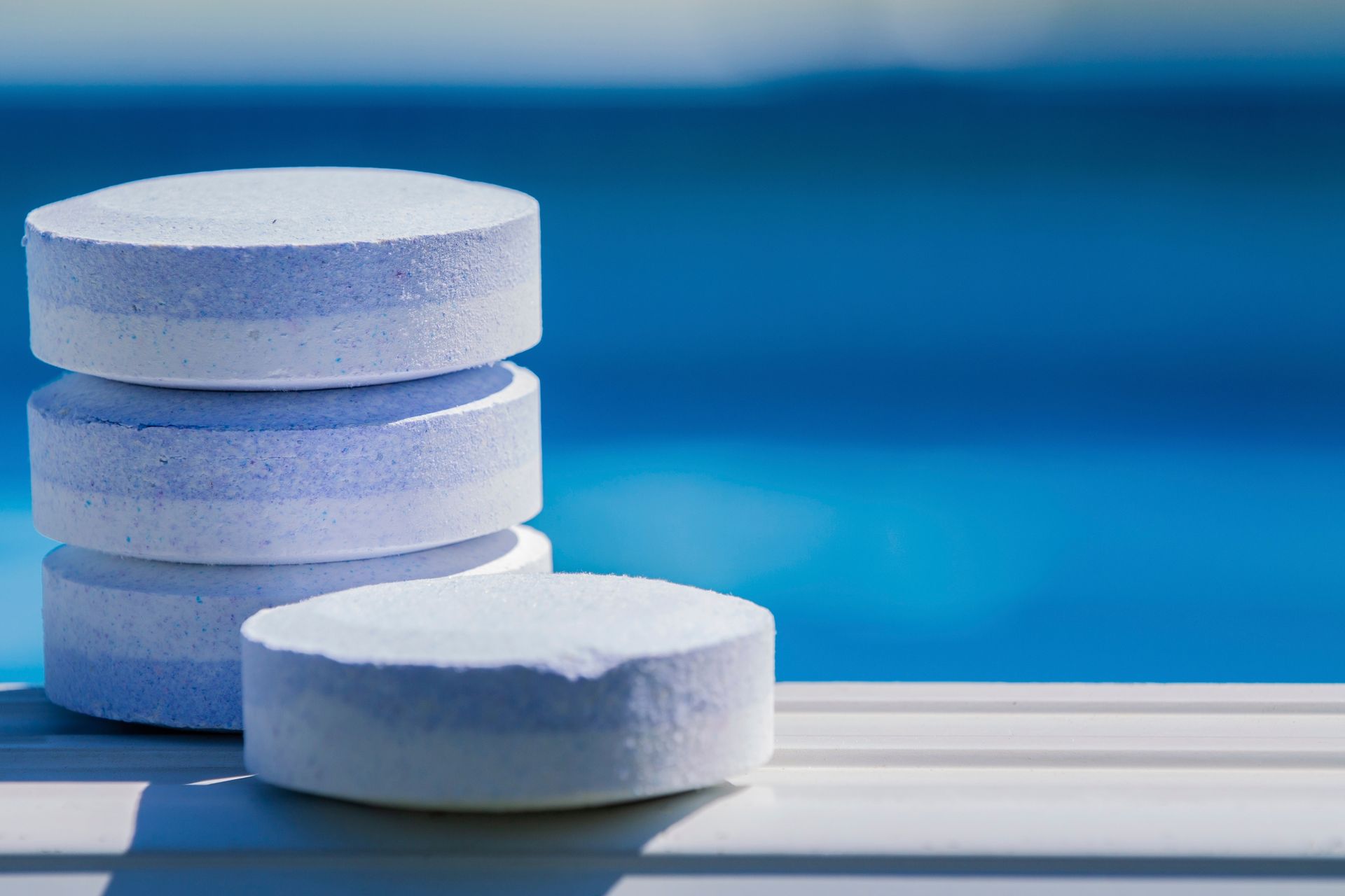 Stack of white pool chlorine tablets against a blue pool.