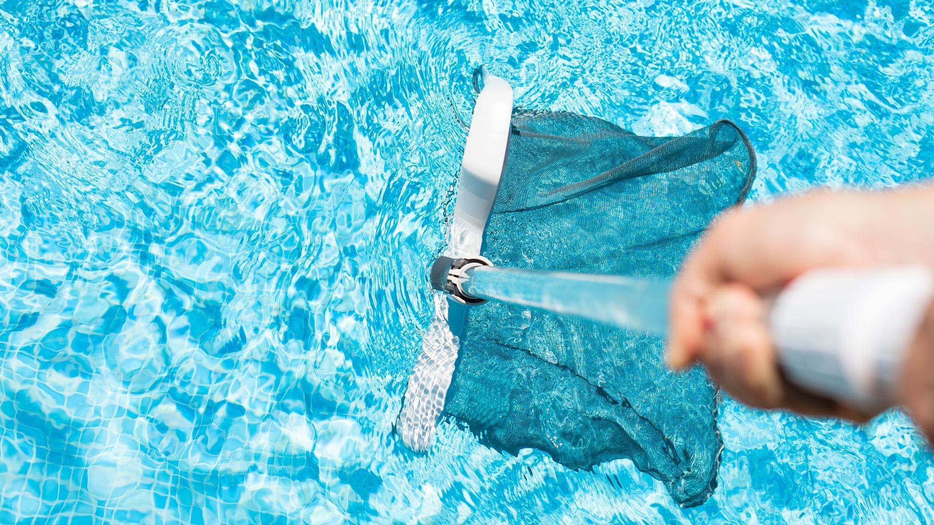 Person uses a pool vacuum to clean a pool's turquoise water; white handle and head.