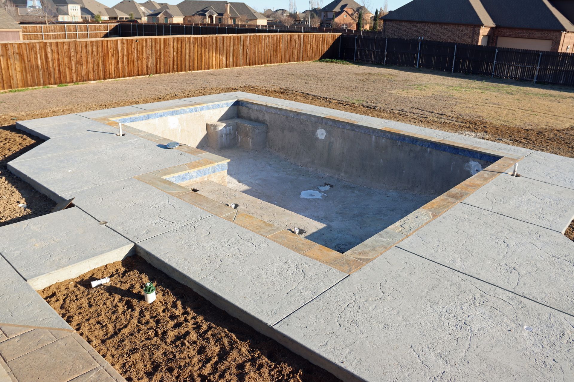 Empty rectangular swimming pool under construction, concrete border, brown fence and yard in background.