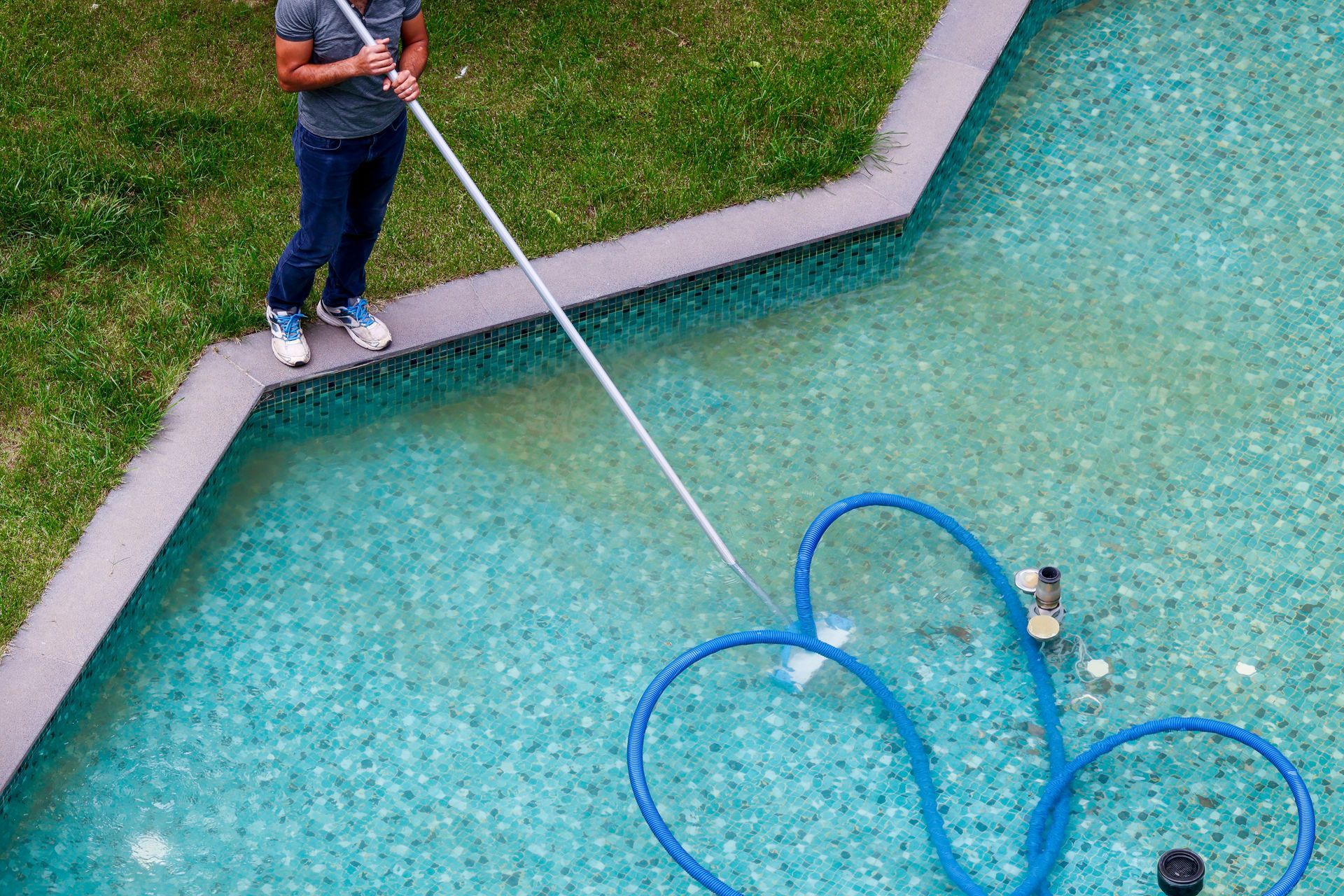 Person cleaning a turquoise pool with a vacuum. Blue hose in the water, green grass nearby.